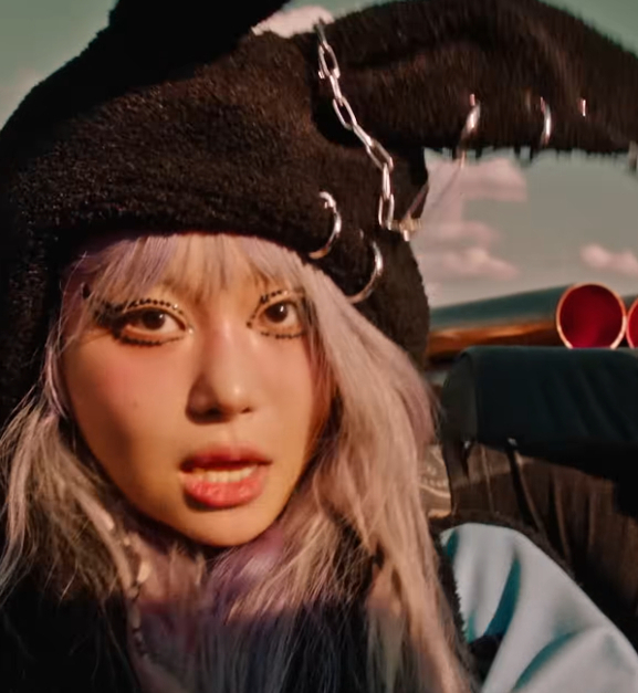 Jvcki Wai with a black bunny ear hat in the backseat of a car. A guy smiling to her right has a buzz cut and tree tattoo on the side of his head.