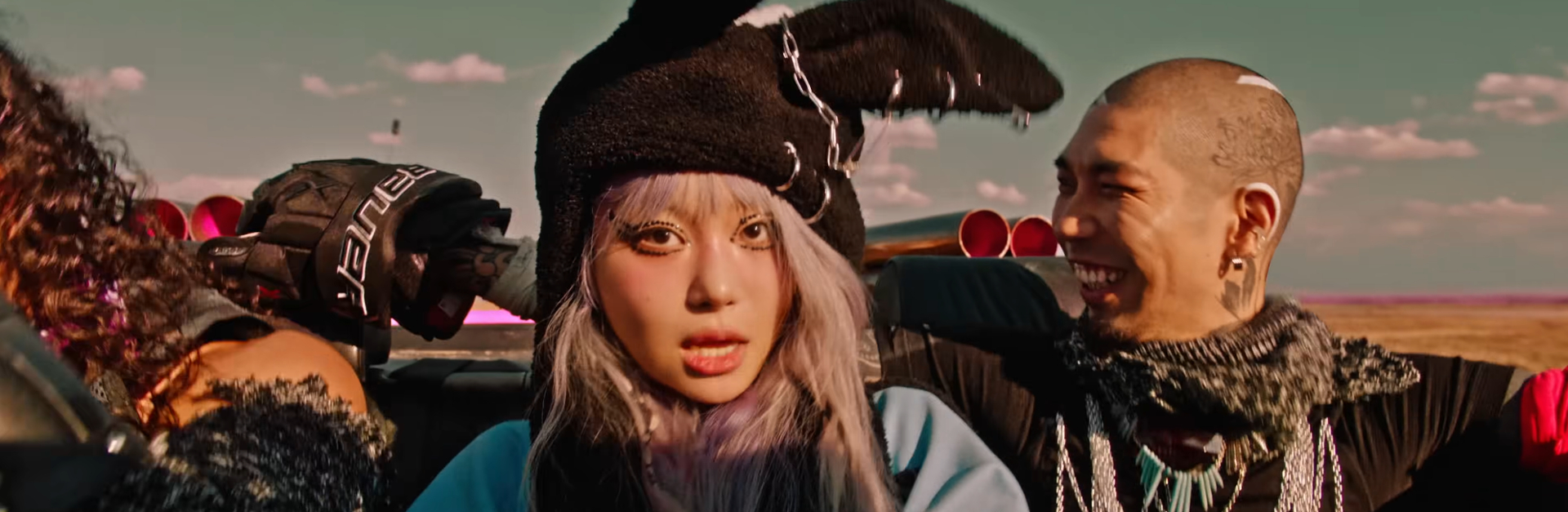 Jvcki Wai with a black bunny ear hat in the backseat of a car. A guy smiling to her right has a buzz cut and tree tattoo on the side of his head.