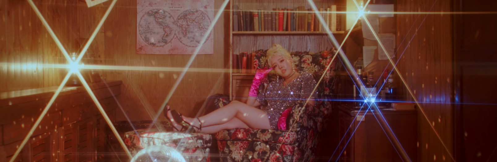 En Sokum sitting in a floral print chair with her legs up on a matching ottoman. The room she is in is wood paneling, along with wood bar, wood bookcase, and wood night stand. There is a disco ball on a table, and light is reflecting with flare off multiple objects.
