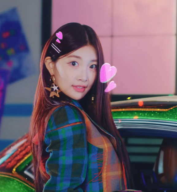 woo!ah! member Minseo is leaning out of a metallic green car window in a garage. She is wearing a flannel jacket, star earrings, and a hairclip. There are digital effect hearts around her head, a sunflower to her left, and colorful stripes on the car.