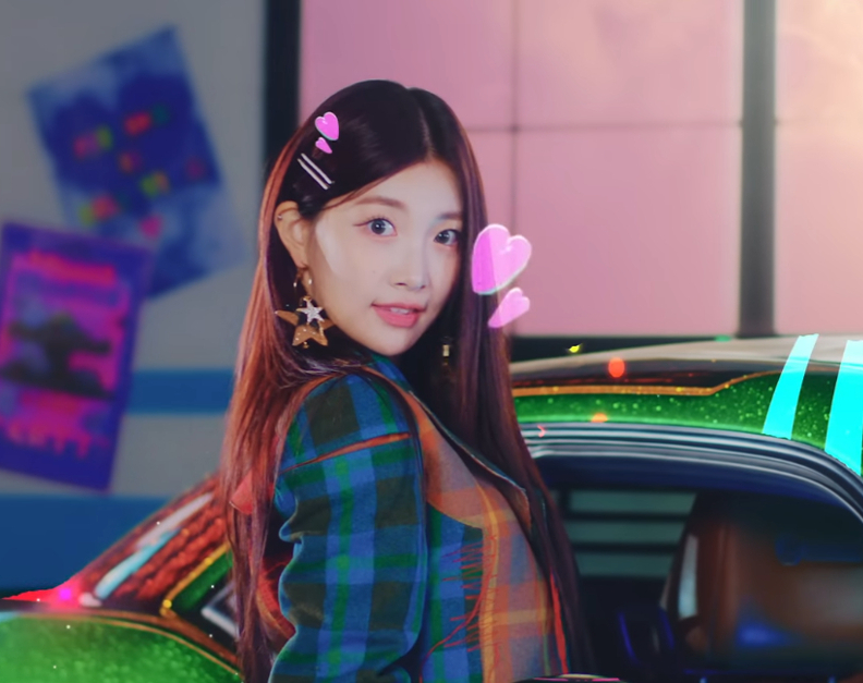 woo!ah! member Minseo is leaning out of a metallic green car window in a garage. She is wearing a flannel jacket, star earrings, and a hairclip. There are digital effect hearts around her head, a sunflower to her left, and colorful stripes on the car.