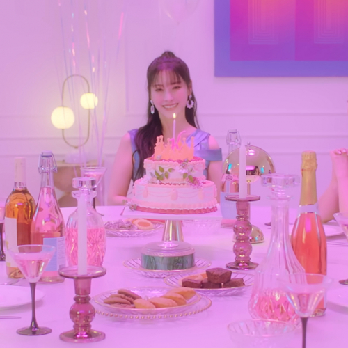 Image shows the 5 members of the girl group KARA sitting around a table smiling. The table has unlit candles, various snacks, plates and silverware, and a cake in the middle with a single lit candle.