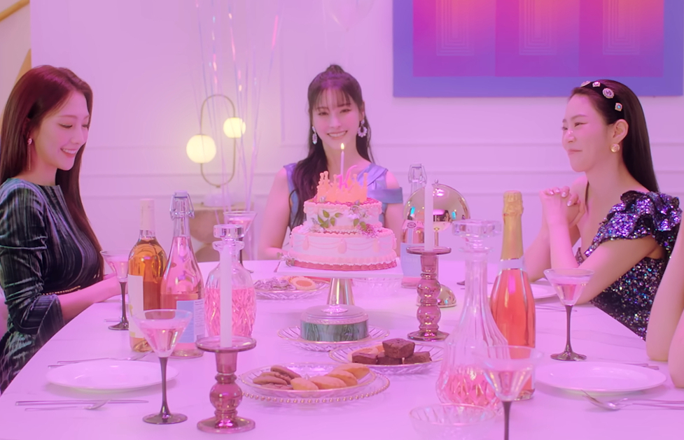 Image shows the 5 members of the girl group KARA sitting around a table smiling. The table has unlit candles, various snacks, plates and silverware, and a cake in the middle with a single lit candle.