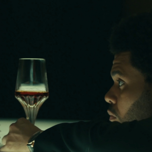 Image shows a black man (The Weeknd) leaning on a counter staring at a glass of liquor