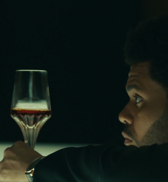 Image shows a black man (The Weeknd) leaning on a counter staring at a glass of liquor