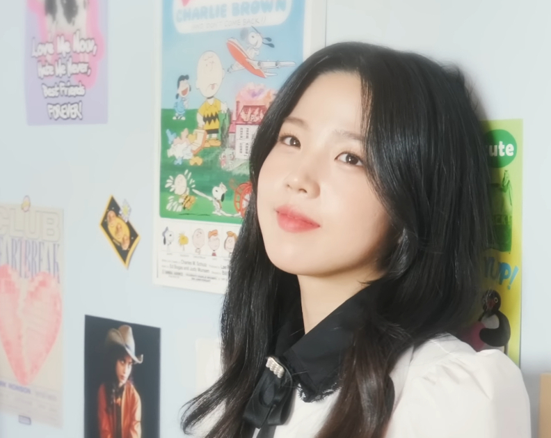 A Korean woman leaves against a wall in a room. The wall has multiple small posters.