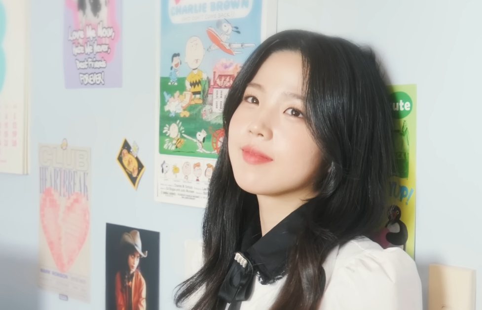 A Korean woman leaves against a wall in a room. The wall has multiple small posters.