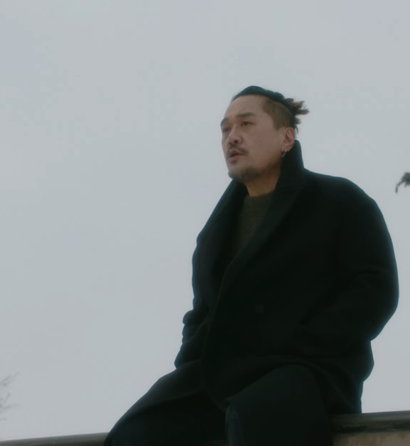 A Korean man sits on a wall wearing a winter coat.