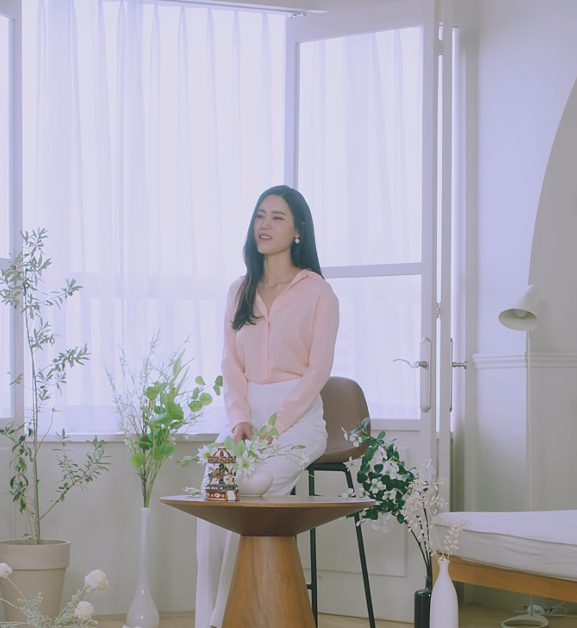 Image shows a Korean woman sitting on a chair in the middle of a room. There are some plants around her and small wooden table in fron of where she is sitting