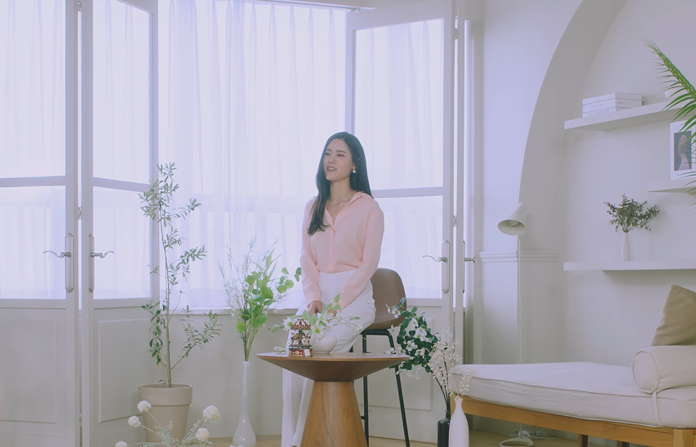 Image shows a Korean woman sitting on a chair in the middle of a room. There are some plants around her and small wooden table in fron of where she is sitting