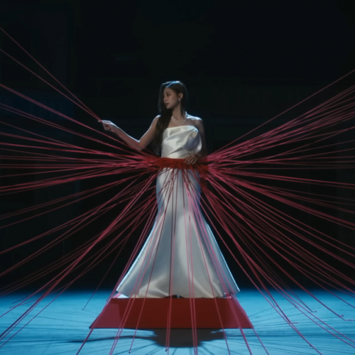 Tzuyu of TWICE is standing on a pedestal with red string wrapped around her waist and spreading out in various directions. She is grabbing on and looking at a string to her right.