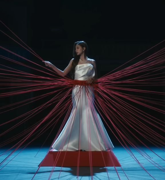 Tzuyu of TWICE is standing on a pedestal with red string wrapped around her waist and spreading out in various directions. She is grabbing on and looking at a string to her right.