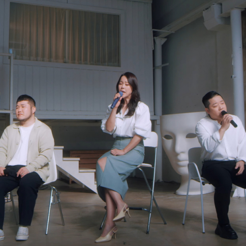 Image shows 2 Korean men and 1 Korean woman sitting in chairs in a large room. The woman and 1 of the men are singing in to mics.