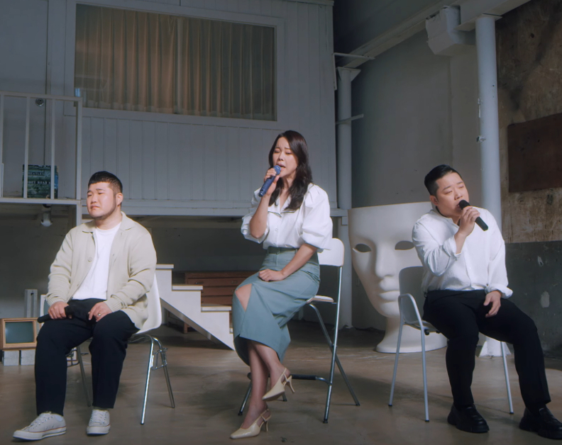 Image shows 2 Korean men and 1 Korean woman sitting in chairs in a large room. The woman and 1 of the men are singing in to mics.