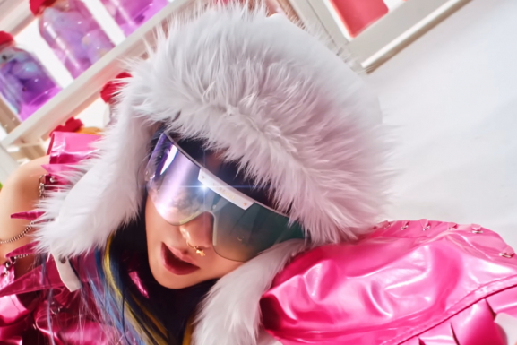 image shows a Korean women laying on a floor. She is wearing a fur hoodie, oversized sunglasses, and a pink vinyl coat