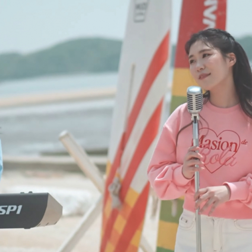 2 Korean women performing on a beach. One is playing a keyboard on the left, and the other standing in front of a microphone stand on the right