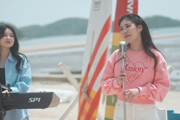 2 Korean women performing on a beach. One is playing a keyboard on the left, and the other standing in front of a microphone stand on the right