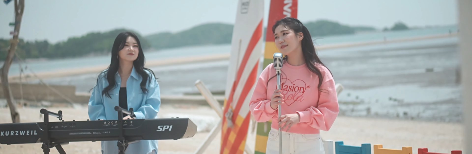 2 Korean women performing on a beach. One is playing a keyboard on the left, and the other standing in front of a microphone stand on the right