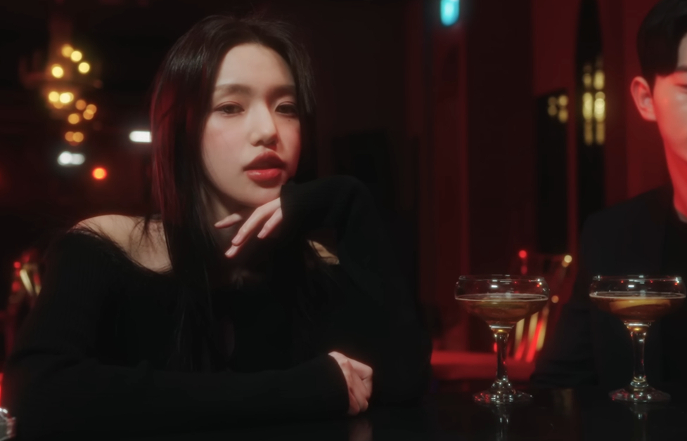 Image shows K-Pop singer iiso sitting at a bar with a man sitting next to her. 2 drinks are on the bar