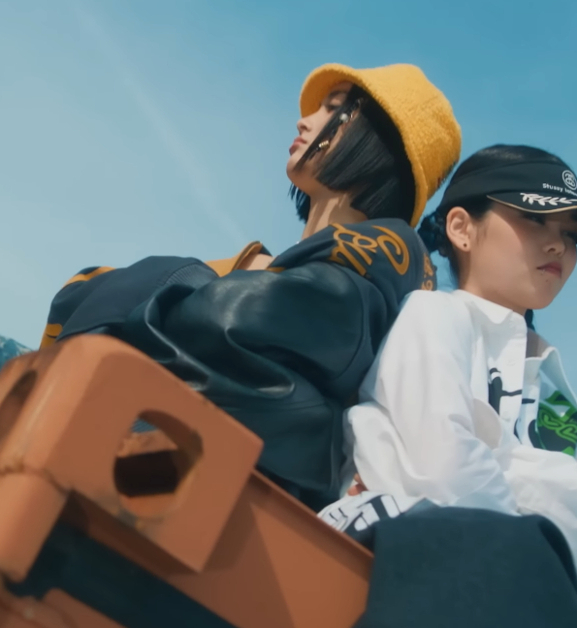 JURIN and COCONA of XG sit back to back on a shipping container with their arms crossed. They are wearing baggy clothes.