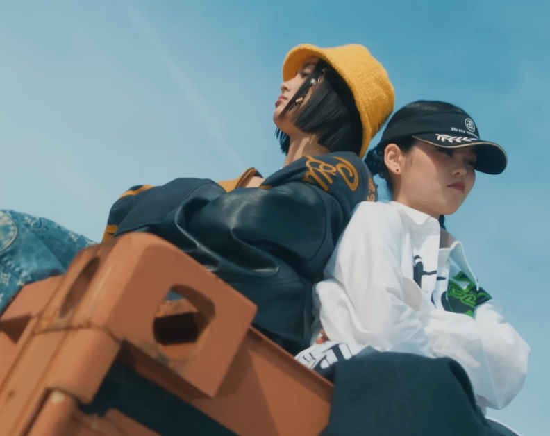 JURIN and COCONA of XG sit back to back on a shipping container with their arms crossed. They are wearing baggy clothes.