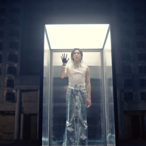 K-Pop singer REN stands in a glass case in front of buildings. He has his right hand pressed against the front glass