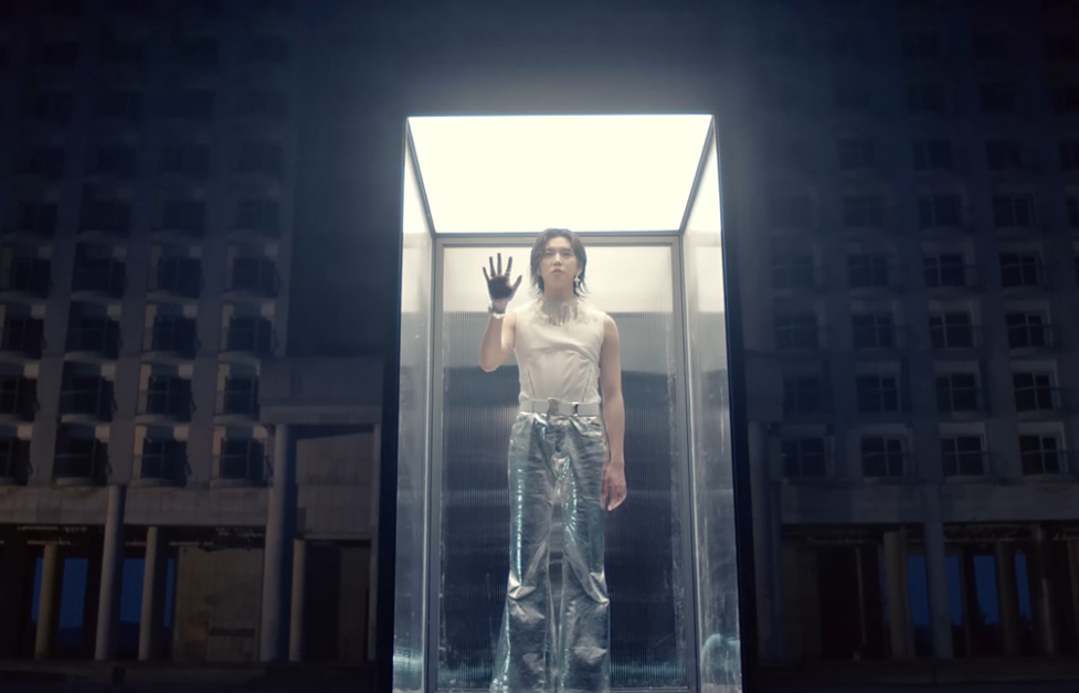 K-Pop singer REN stands in a glass case in front of buildings. He has his right hand pressed against the front glass