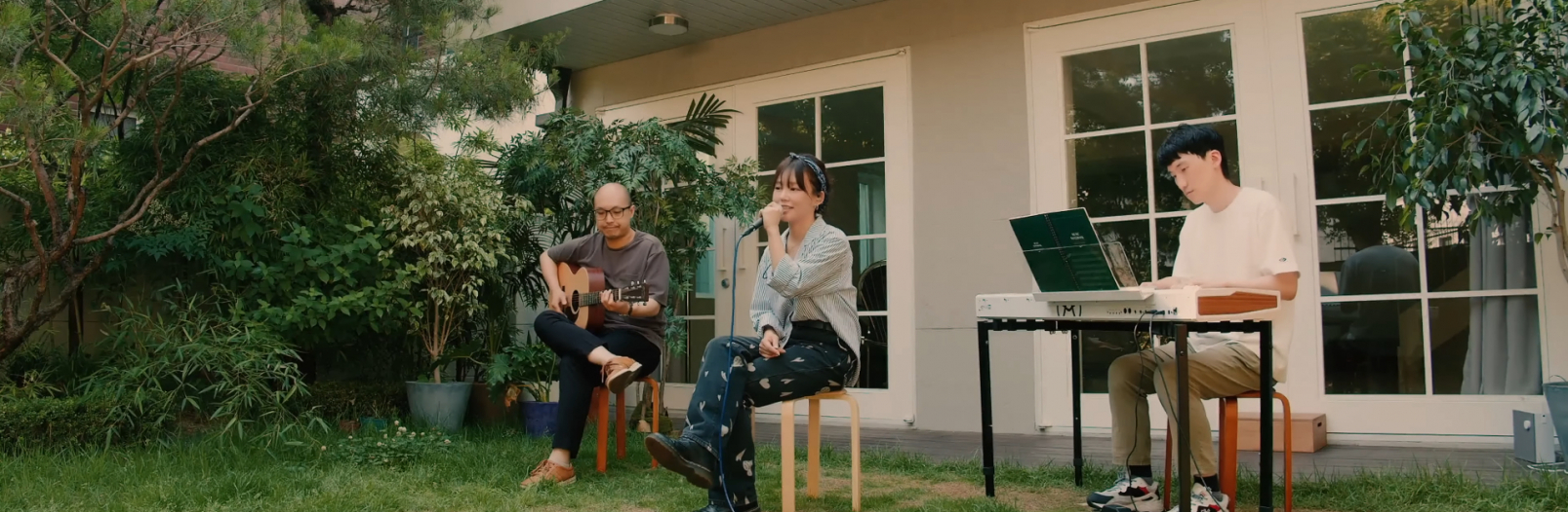 DONNA and 2 Korean men are sitting in a yard behind a building. Donna is singing in to a microphone. The man to her right is playing guitar and the one to her left playing the keyboard.