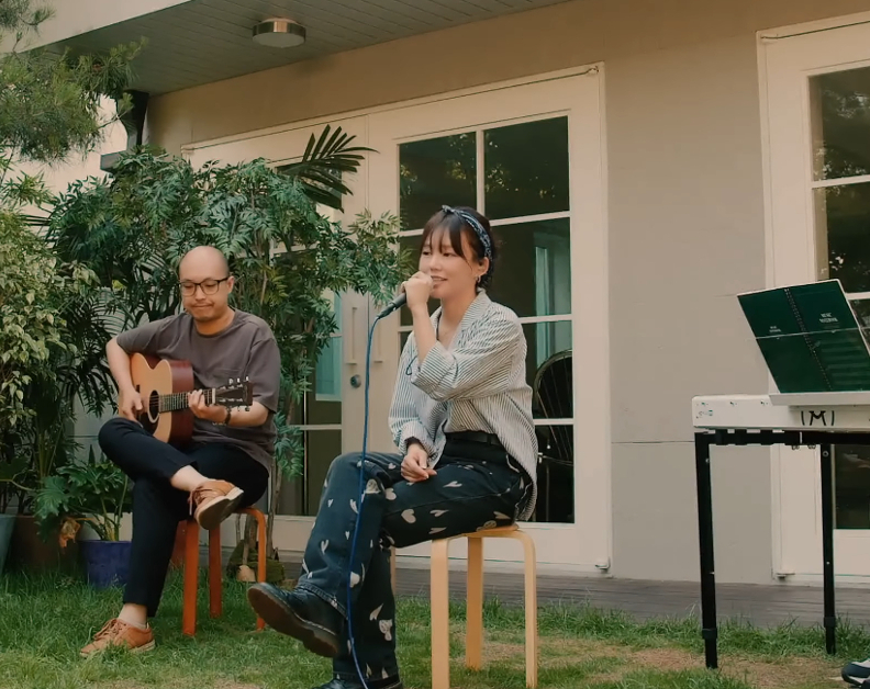 DONNA and 2 Korean men are sitting in a yard behind a building. Donna is singing in to a microphone. The man to her right is playing guitar and the one to her left playing the keyboard.