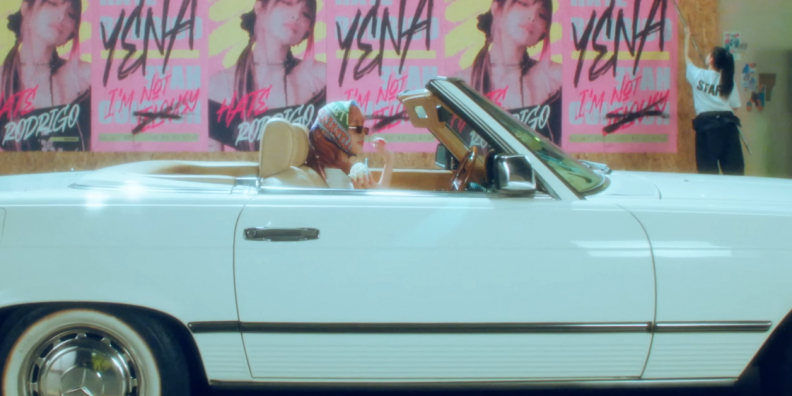 K-Pop singer Yena sits in a convertible holding ice cream. Promotional posters are on the wall behind them