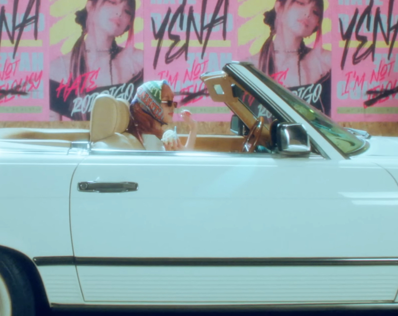 K-Pop singer Yena sits in a convertible holding ice cream. Promotional posters are on the wall behind them