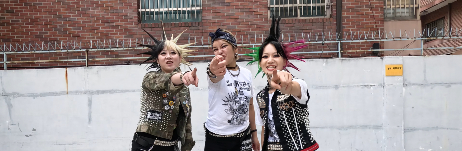 3 members of RUMKICKS are pointing at the camera. They are all dressed up in punk rock gear. The members on the left and right have spiked hair, while the middle member is wearing a bandana around her head that is tied in the front.