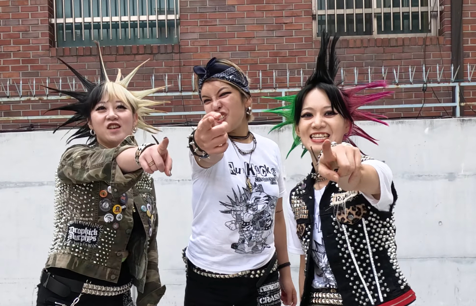 3 members of RUMKICKS are pointing at the camera. They are all dressed up in punk rock gear. The members on the left and right have spiked hair, while the middle member is wearing a bandana around her head that is tied in the front.