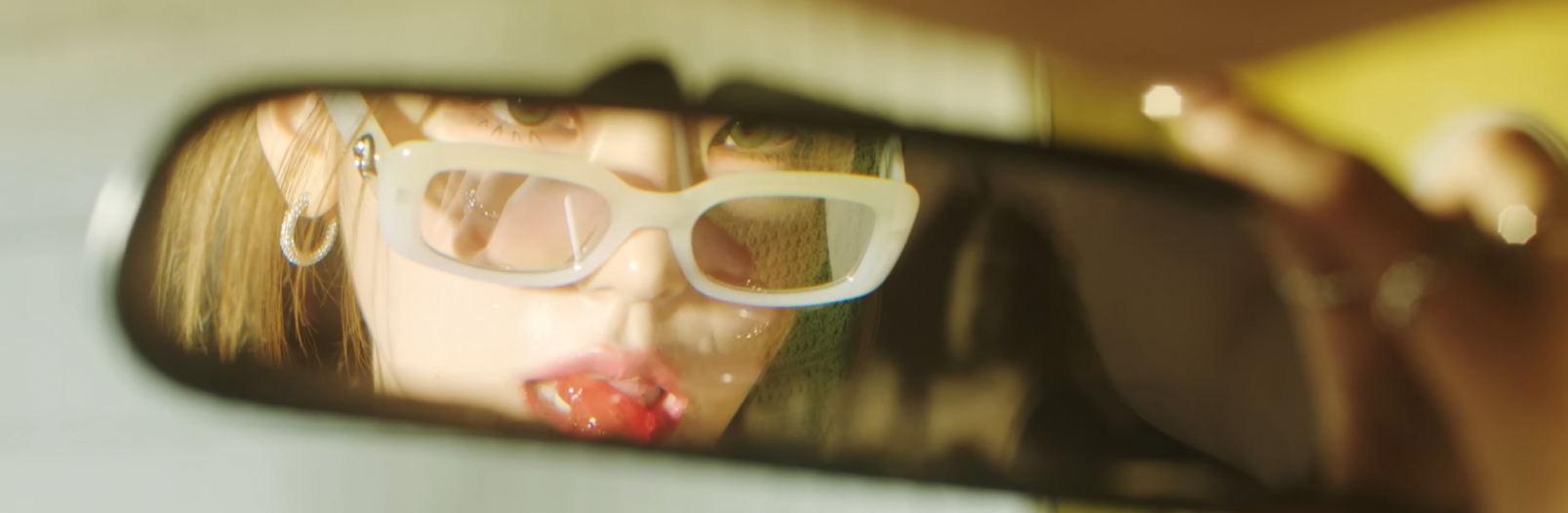 In a truck's rear view mirror we see a partial reflection of Yeji from ITZY. We see part of her face, and she is wearing glasses down on the tip of her nose as she looks in the mirror and puts a lollipop to her mouth
