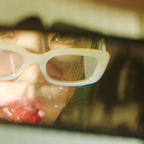 In a truck's rear view mirror we see a partial reflection of Yeji from ITZY. We see part of her face, and she is wearing glasses down on the tip of her nose as she looks in the mirror and puts a lollipop to her mouth