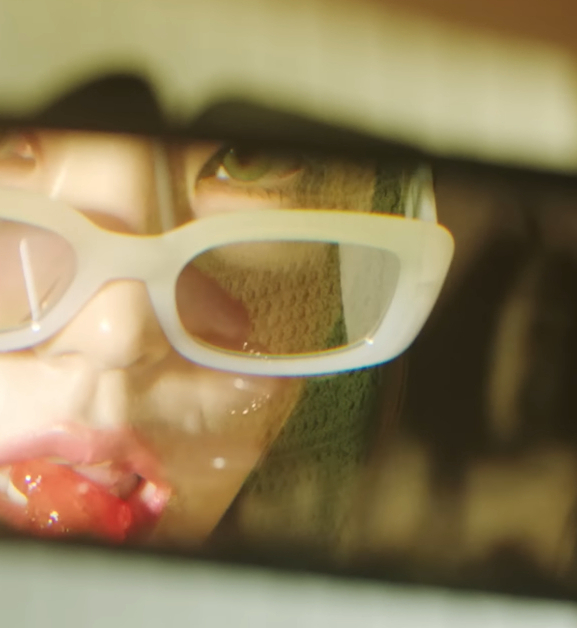 In a truck's rear view mirror we see a partial reflection of Yeji from ITZY. We see part of her face, and she is wearing glasses down on the tip of her nose as she looks in the mirror and puts a lollipop to her mouth