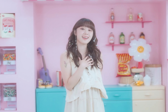 K-Pop singer, Woo Yerin, stands in front of a cabinet setup. The counter features various foods, and knick knacks. There are shelves filled with drinks and snacks.