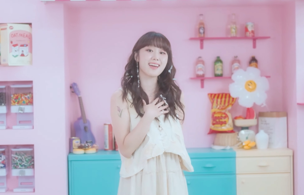 K-Pop singer, Woo Yerin, stands in front of a cabinet setup. The counter features various foods, and knick knacks. There are shelves filled with drinks and snacks.