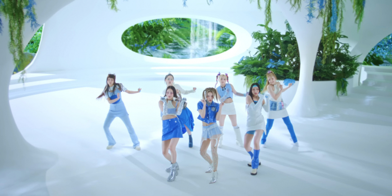 7 members of girl group, XG, are dancing in an all white building. You can see through open spaces fauna and water in the background.