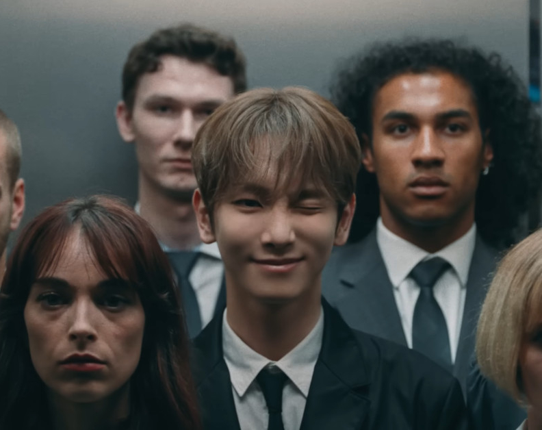 Singer KEY is standing in a crowded elevator winking at the camera