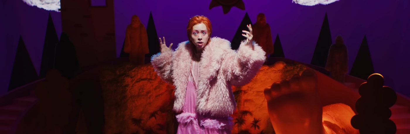 Zior Park is standing elevated in front of a stage set. He is holding his arms out, while wearing a pink fur coat and a very long pink dress.