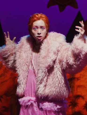 Zior Park is standing elevated in front of a stage set. He is holding his arms out, while wearing a pink fur coat and a very long pink dress.