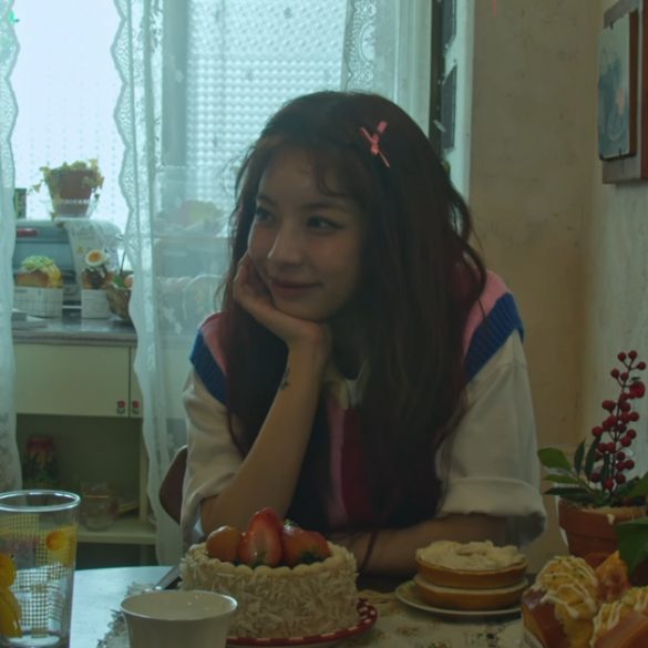 Juniel is sitting at a table in a kitchen. She is resting her chin on her palm. On the table is a strawberry cake, a glass, a mug, bagel w/ creamcheese, and 2 breakfast sandwiches.