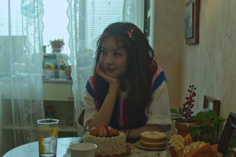 Juniel is sitting at a table in a kitchen. She is resting her chin on her palm. On the table is a strawberry cake, a glass, a mug, bagel w/ creamcheese, and 2 breakfast sandwiches.