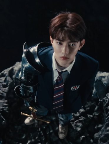 A member of boy group, ZEROBASEONE is standing on a small rock in a body of water holding a sword and wearing a school uniform. He is looking up at the camera