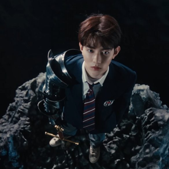 A member of boy group, ZEROBASEONE is standing on a small rock in a body of water holding a sword and wearing a school uniform. He is looking up at the camera