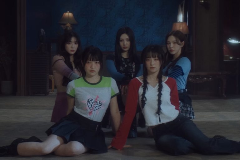 5 members of Red Velvet are sitting and kneeling together in a dimly lit room