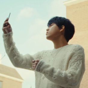 Lee Mu Jin is standing outside holding up a flip cell phone
