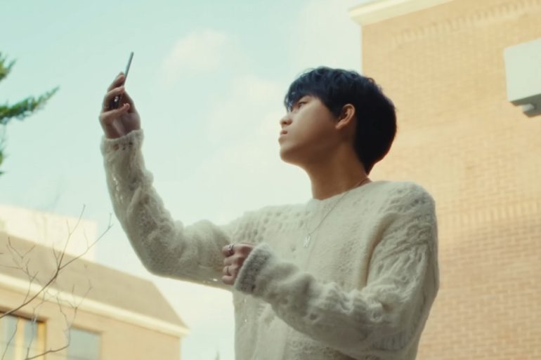 Lee Mu Jin is standing outside holding up a flip cell phone