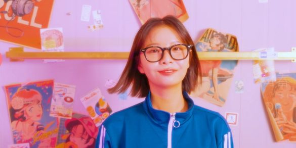 MiJoo is standing in front of a wall wearing glasses and a track suit top. Behind her on the wall are stickers, posters featuring anime girls, overalls, and a mic cord.